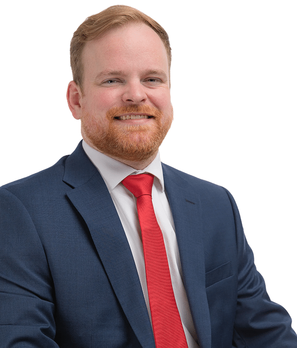 A man with short red hair and a full beard, wearing a dark suit, white shirt, and bright red tie, poses against a plain white background.