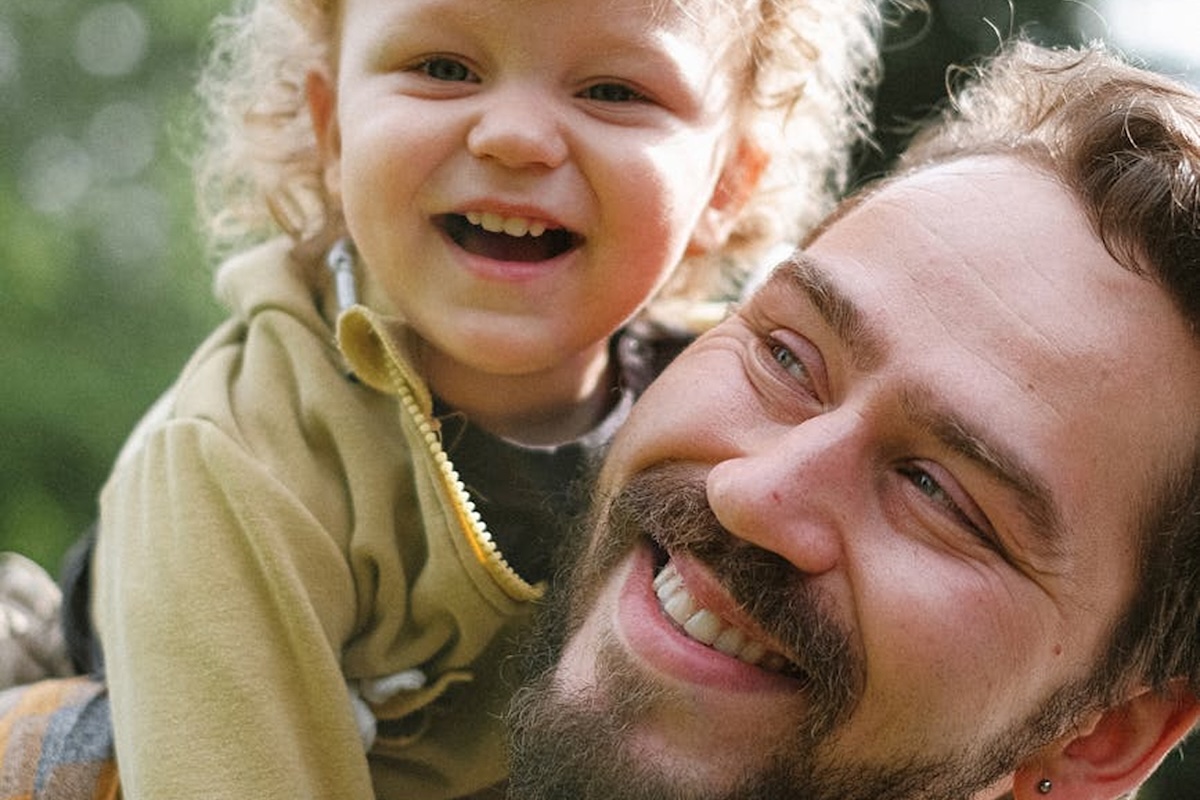 A smiling child with curly hair is being held by a bearded man, both looking happy and joyful outdoors. The background is blurred greenery.