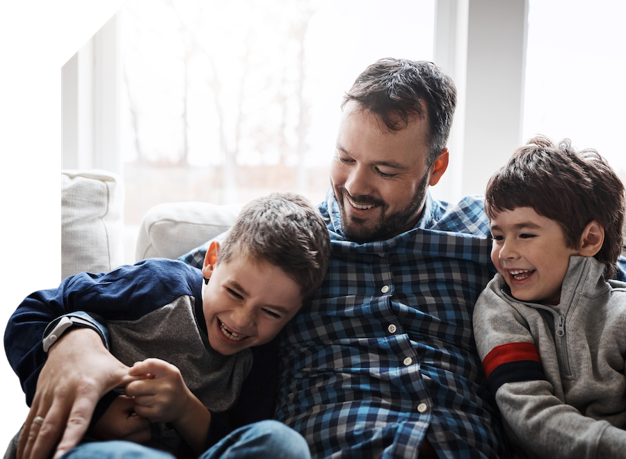 A man sits on a couch smiling with two young boys, who are laughing and leaning close to him. They appear happy and relaxed in a bright, cozy living room.