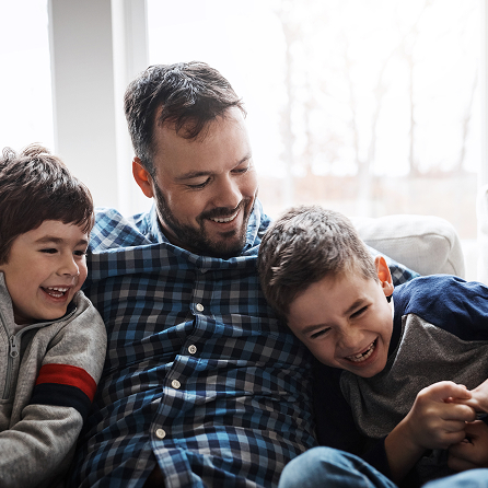 A man sits on a couch smiling with two young boys, who are laughing and leaning close to him. They appear happy and relaxed in a bright, cozy living room.