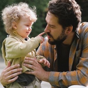 A young child with curly hair touches the nose of a bearded man wearing a plaid shirt. They are smiling and making eye contact outdoors, sharing a playful and affectionate moment.