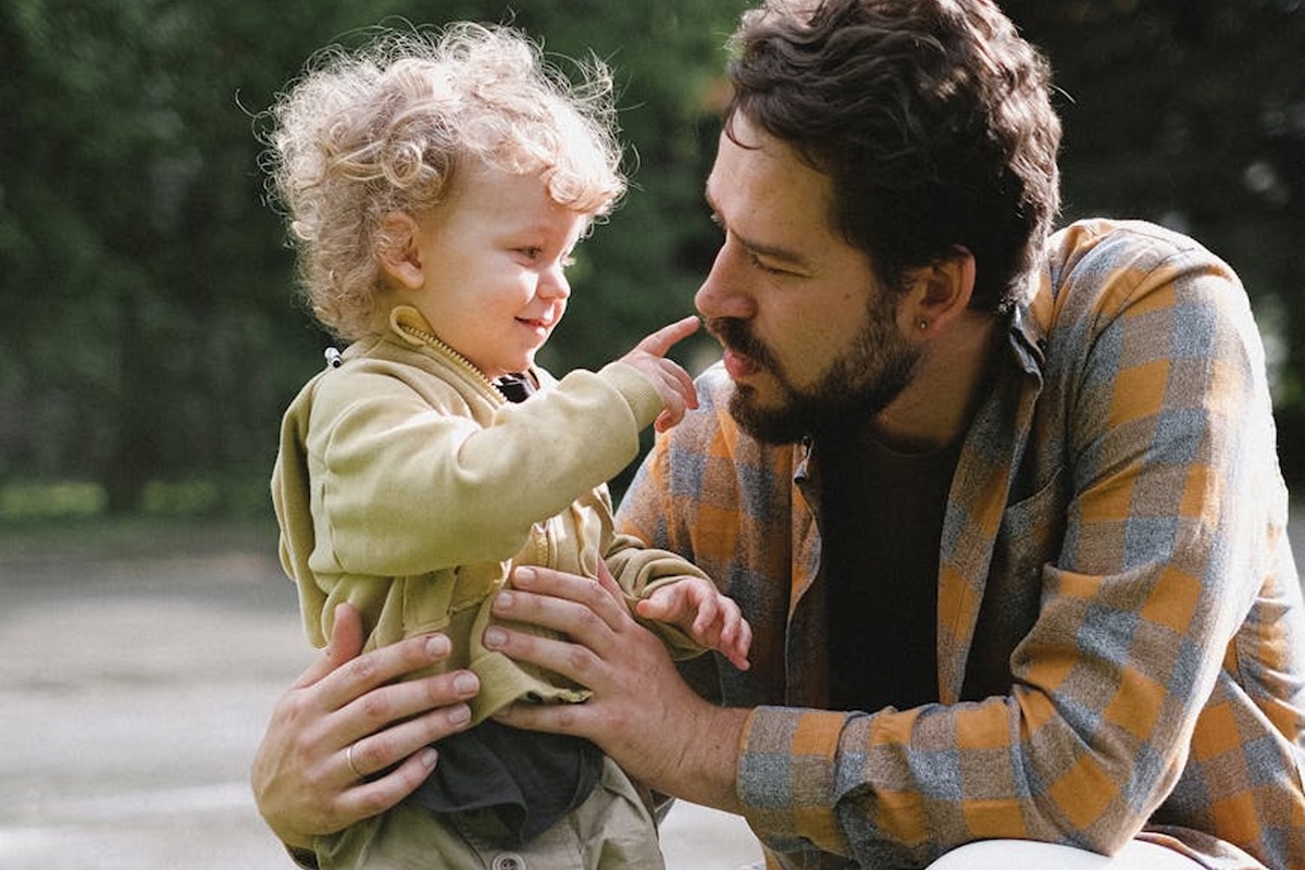 A man with a beard crouches down and smiles as a young child with curly hair touches his nose outdoors, both wearing casual jackets.